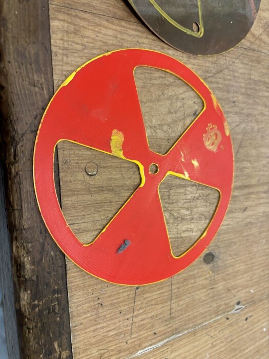 Röd, rund metallskiva med tre spjällöppningar, kanske en ventilationsdel, ligger på ett träbord.