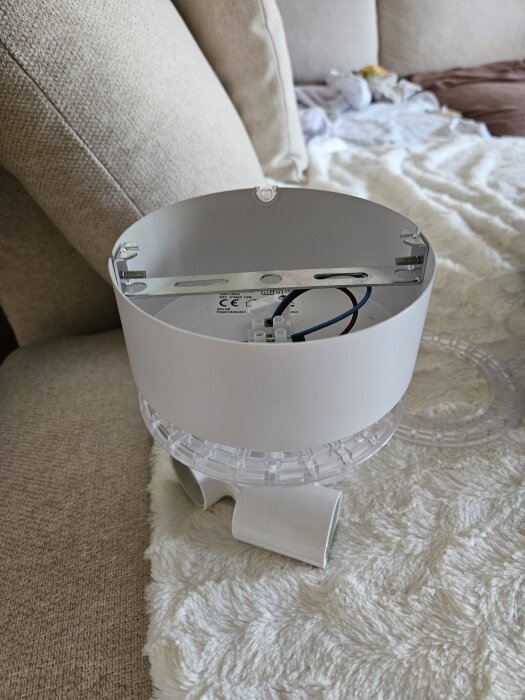 Vit lampsockel med synliga kablar och monteringsfäste placerad på beige soffa.
