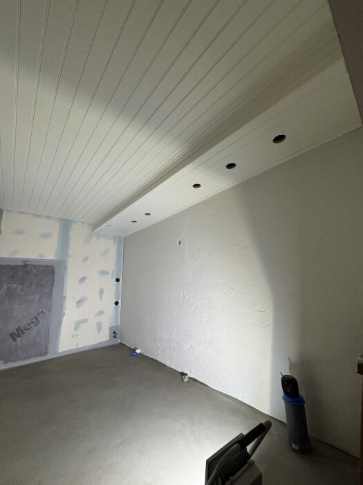 Vitmålat rum under renovering med spottar i taket och oavslutad vägg. Verktyg och material synliga på golvet.