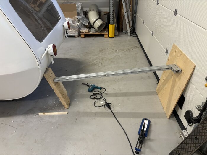 Släpfordon i garage med improviserat stöd i trä och metall, omgiven av verktyg och byggmaterial.
