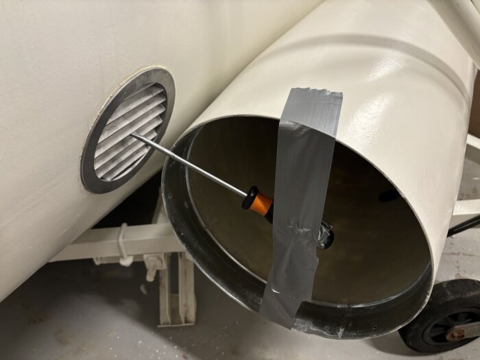 Aluminiumventil med galler och skruvverktyg fäst med silvertejp på vit metallkonstruktion för bastuventilation.