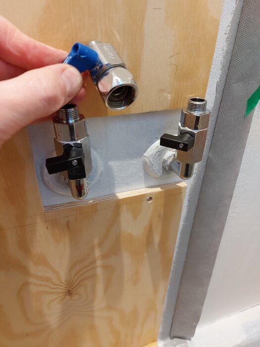 En hand håller en kulventil ovanför två monterade kulventiler på en plywoodvägg, del av ett badrumsrenoveringsprojekt.