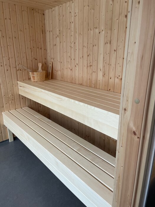 Bastuinteriör i trä med två bänkar, bastukorg och skopa placerade på den övre bänken.