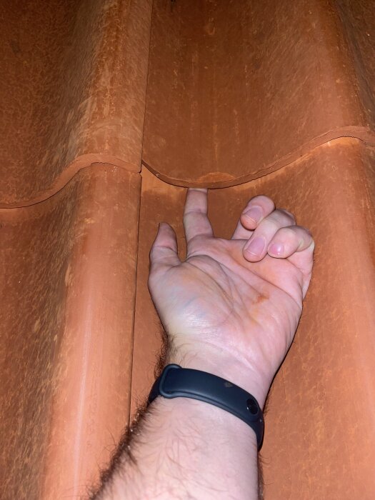 En hand pekar på en glipa mellan enkupiga tegelpannor på ett tak.