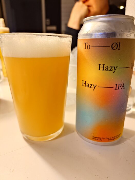 Ett glas med skummig, oklar öl och en färgglad burk To ØL Hazy IPA på ett bord inomhus.