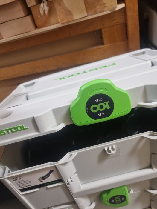 Festool systainer med grön låsmekanism som inte går att öppna helt, placerad framför en hylla med träblock i bakgrunden.