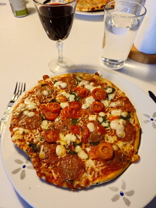 Pizza med pepperoni, tomatskivor och ost på en tallrik, bredvid ett glas rött vin och ett glas vatten.