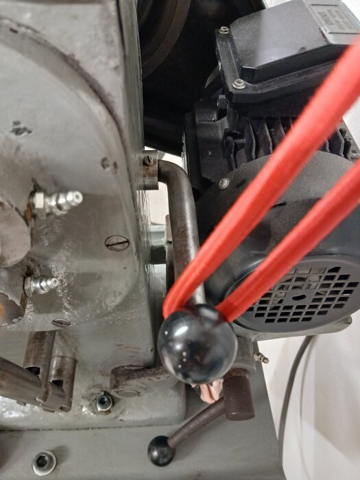 Maskindel med en spak som hålls uppe av ett rött snöre, bredvid en motor; frågeställning om hur man bäst stödjer spaken.