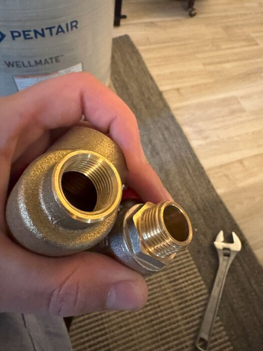 Hand håller i två metallkopplingar till VVS-system, en med invändig och en med utvändig gänga, verktyg i bakgrunden.