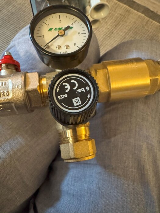 Tryckmätare och ventildel placerad på en tygbakgrund, med en tryckskala från 0 till 6 bar och koppling i metall.