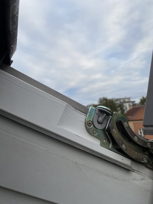 Metallplatta och gångjärn vid skarv mellan fönsterkarm och fönsteröppning på ett Velux GGL CO2-fönster, med utsikt mot himmel och hus i bakgrunden.