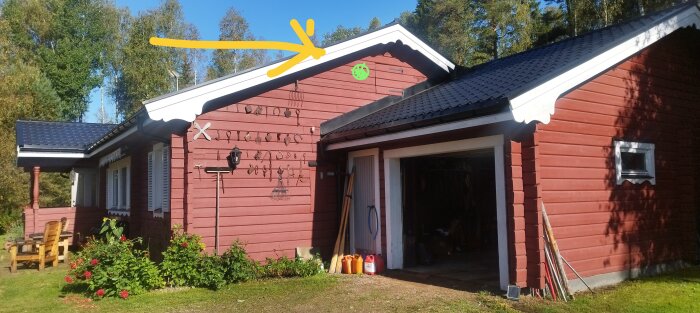 Röd träbyggnad med svart tak och en gul pil som pekar mot takutsprånget. Olika verktyg hänger på väggen. Grön markering bredvid pilen.