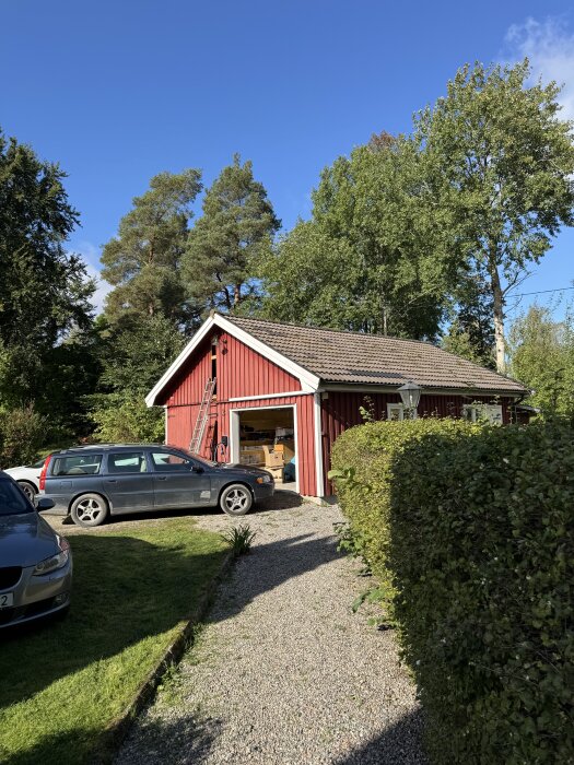 Rött garage med bil utanför, stege lutad mot väggen. Bredvid finns en carport. Träd och buskar omger området.
