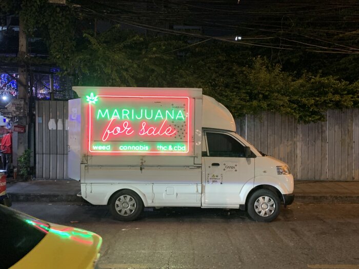 Vit skåpbil med neonljusskylt "Marijuana for sale" parkerad på gata i Thailand, upplyst på natten.