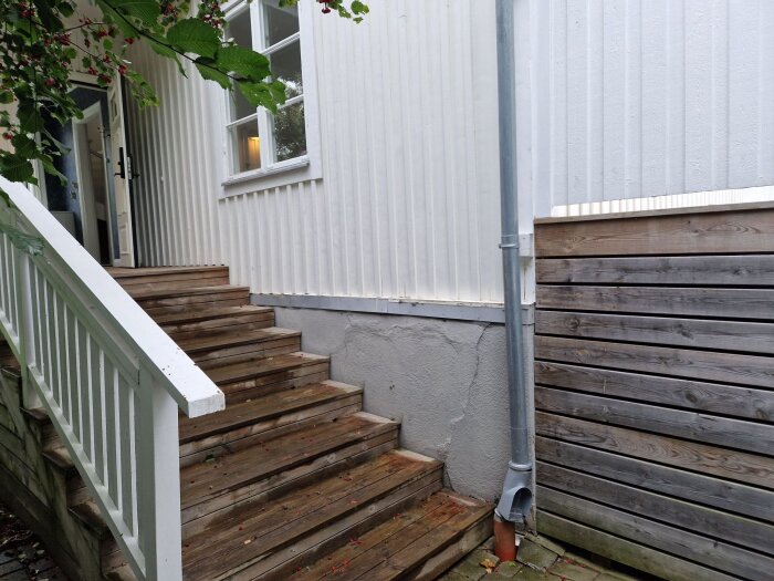 Trappa med vit ledstång vid hus med sprickor i sockeln, dräneringsrör och träpanel.