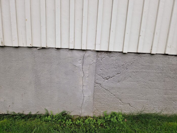 Spricka i en husgrund med lagningar och synliga tecken på tidigare reparationer, intill ett vitt träfasadområde.