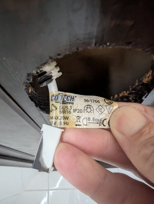 Bränd kabel nära takspot med etikett ”COLTECH LIGHTING” och specifikationer synliga; handen håller i kabeln med skadad isolering.