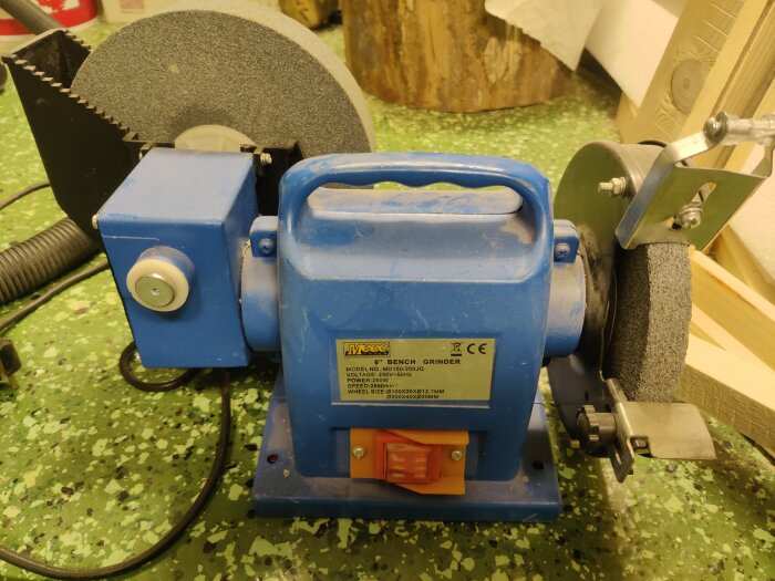 Blå bänkslipmaskin med två slipstenar, en stor och en liten, utan synligt anhåll, placerad på grön golvyta.