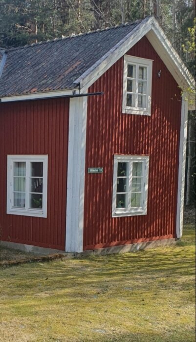 Rött hus med vita fönsterramar och grön bakgrund med vit text på en skylt vid väggen.