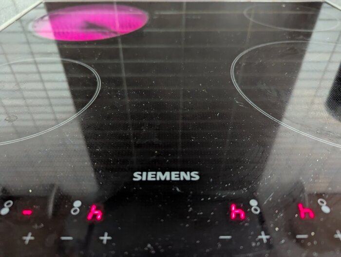Keramisk spishäll med varningslampor tända, en platta längst in till vänster är aktiv med röd glöd. Märkt med "Siemens".