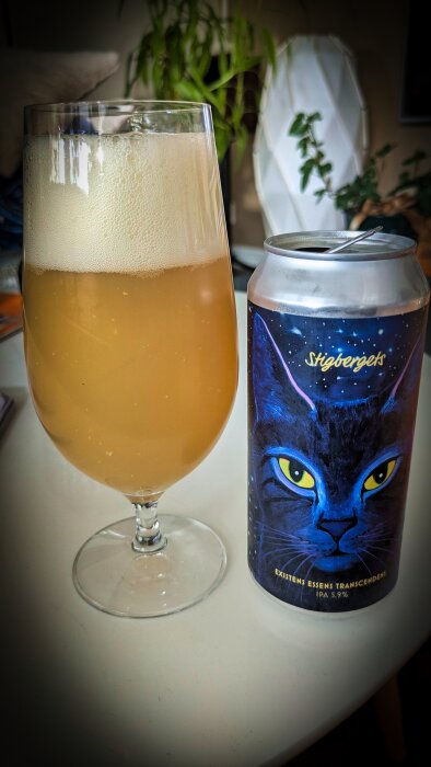 Glas med öl och en ölburk med en illustration av en katt på etiketten, stående på ett bord.