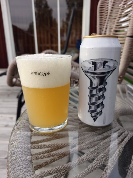 Ett glas ljus öl med skumkrona bredvid en burk med en skruvmotiv, stående på ett glasbord.