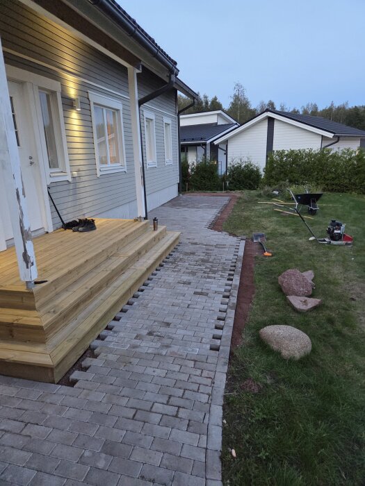 Trätrappa och nylagd stenläggning vid husentré, omgivna av gräs och verktyg som spade och skottkärra på sidan.