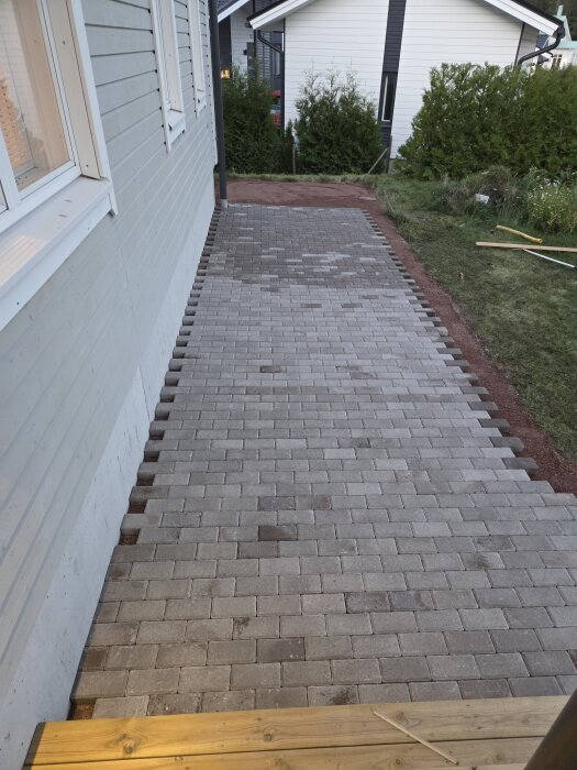 Nylagd stenläggning längs en husvägg med en klar gångväg av gråa plattor; lantligt område i bakgrund.