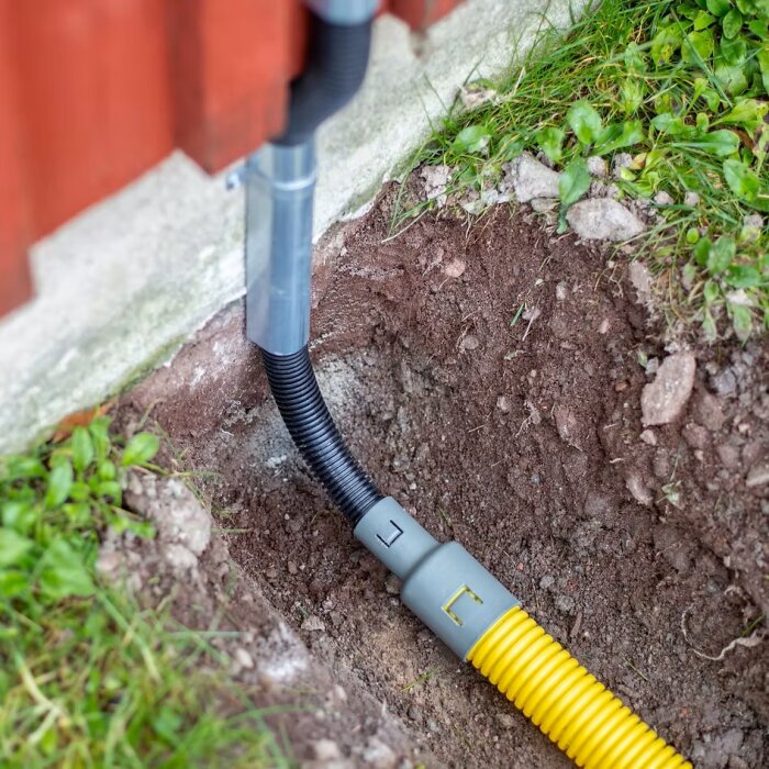 Markrör med övergång till flexslang nära husvägg, inuti ett grunt hål med synlig jord och gräs.