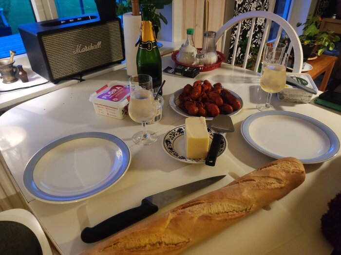 Matbord med ost, bröd, kräftor, vinglas och smörpaket. En flaska champagne syns i bakgrunden, och en Marshall-högtalare står på fönsterbrädan.