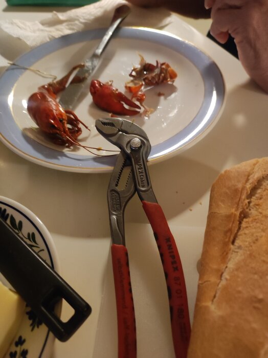 En knäcktång ligger på ett bord bredvid en tallrik med kräftor och ett bröd.