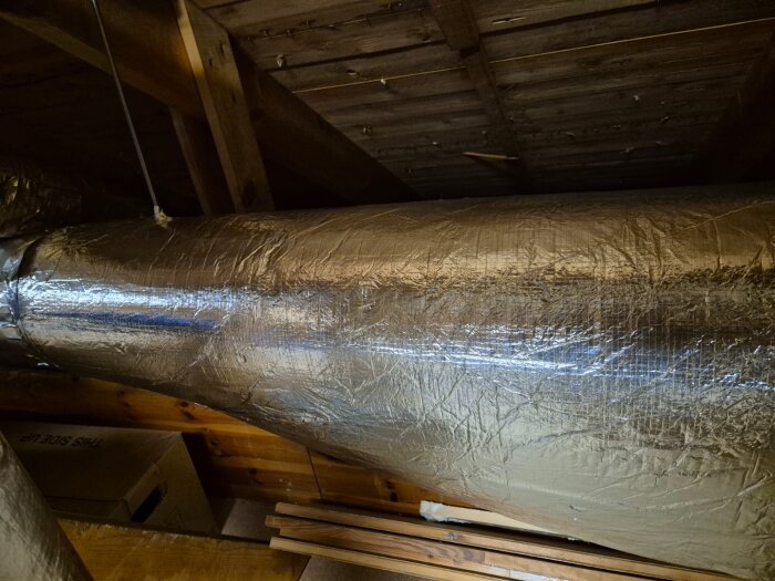 Isolerad ventilationskanal i vindsutrymme med trätak och kartonger.