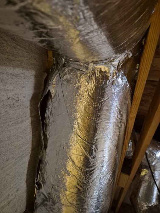 Isolerade rör lindade med aluminiumfolie i en byggnad, monterade nära betongvägg och träkonstruktion.