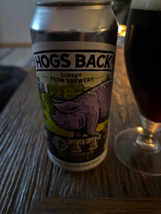 Burk med Hogs Back öl, etikett med lila gris och text "Surrey Farm Brewery". Glas med mörk öl bredvid.