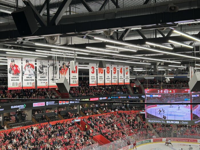 Hockeyarena med hängande spelarbanners i taket, fullsatt med rödklädd publik, stor skärm visar pågående hockeymatch.