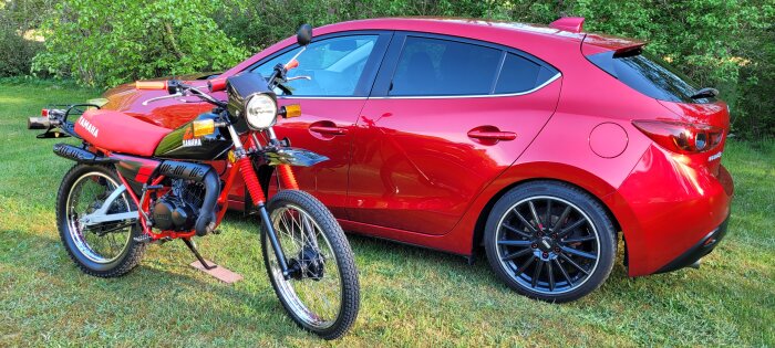 Röd bil och röd Yamaha-motorcykel på gräsmatta med grönskande bakgrund.