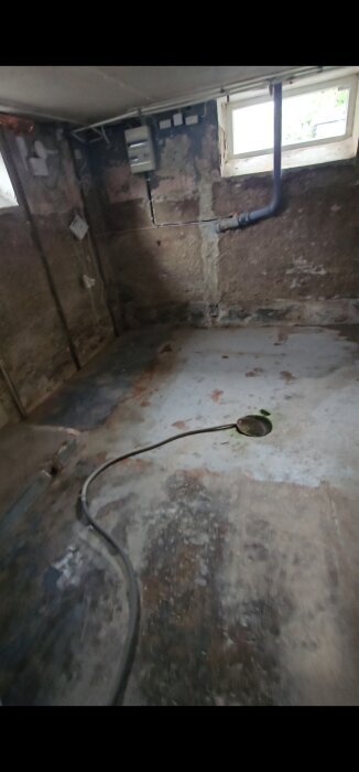 Gammalt källarrum med rör på vägg och golv, ett fönster upptill, och en betongyta med ett öppet hål i mitten där rör eller kablar syns.