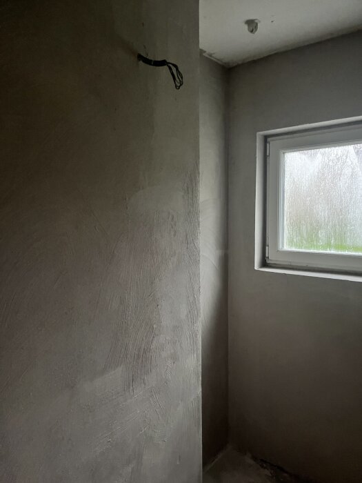 Ofärdig badrumsnisch med fönster, omålad putsad vägg och en svart kabel hängande från taket.