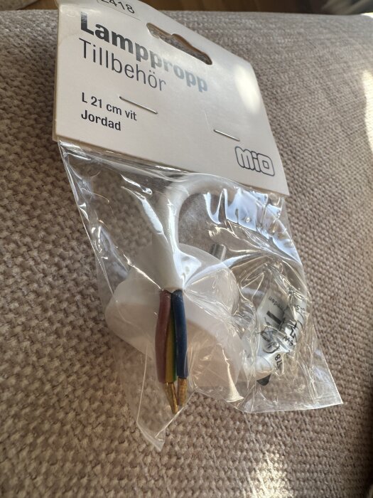 Förpackning med en vit, jordad lamppropp från Mio, modell L 21 cm, synlig med kopparändar i tre färger: brun, blå och gul-grön.