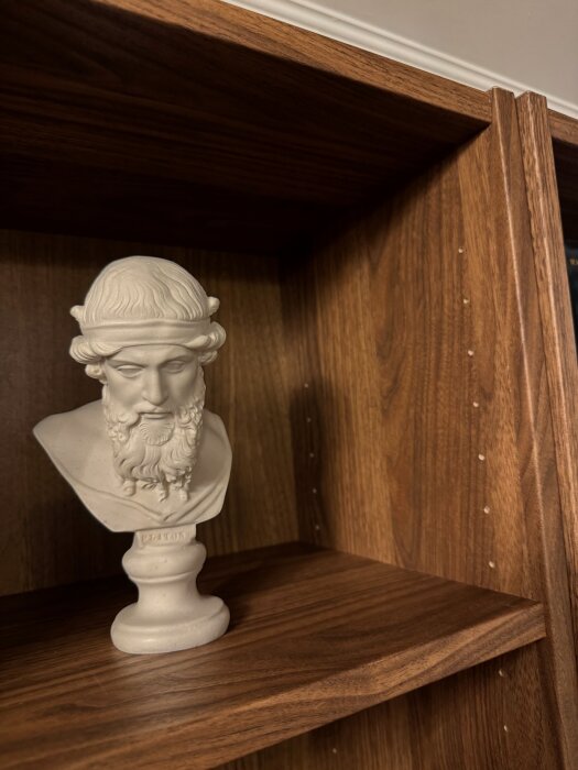 Brun bokhylla med två hyllplan och en skulptur av en byst, eventuellt av den grekiska filosofen Platon, placerad på det nedre hyllplanet.