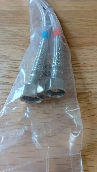 Två flexibla metallslangar med blå och röd markering, förpackade i plast, ligger på ett trägolv.