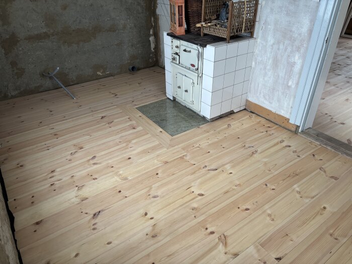 Kök med trägolv och gammal eldstad, synlig avloppsrör mot en lerklädd vägg, förberedelser för att regla upp väggen.