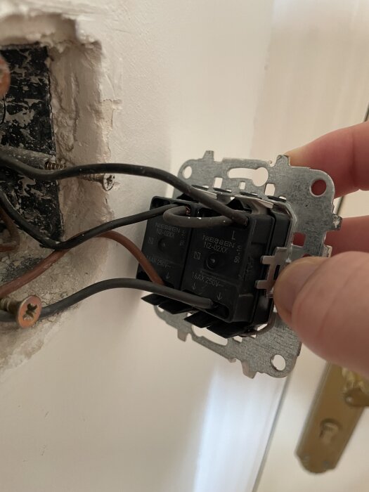 En hand håller i en svart dubbeldimmer kopplad till elkablar i ett vägguttag, redo för installation eller byte av strömbrytare.