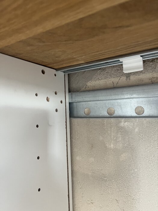 Bild av undersidan av köksskåp, visar träpanel, metallbeslag och en rad borrhål, används i diskussion om att montera ut ett skåp för diskmaskin.