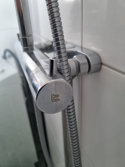 Kromfärgad duschblandare med en vridbar temperaturkontroll och ansluten duschslang syns nära kaklad vägg.