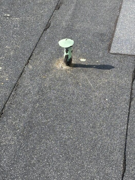 Ventilationshuv på ett låglutande papptak, omgiven av asfaltbeläggning, med tecken på slitage och en grön hattliknande topp.