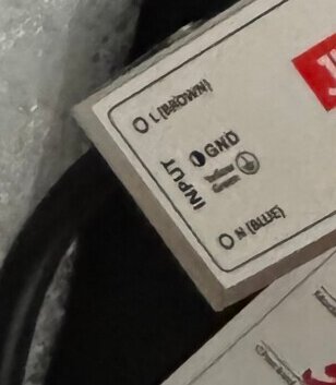 Etikett med jordningssymbol och anslutningsinformation på elektronisk enhet; markering för brun, blå och jordledare.