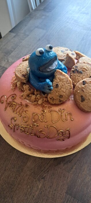 Rosa tårta med texten "Grattis på Din Grattis Dag", dekorerad med figur av blå figur som håller chokladkakor.