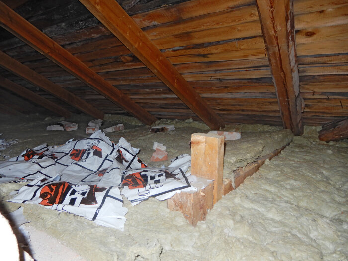 Vindsutrymme med ohyvlat virke i taket, ingen fuktspärr och synliga isoleringsmaterial och murstenar.