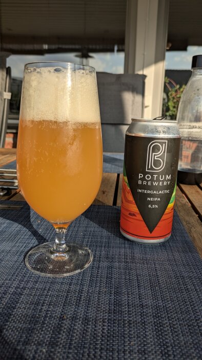 Ölglas fyllt med grumlig, gyllene öl bredvid en burk med etikett "Potum Brewery Intergalactic NEIPA 6,5%" på ett bord utomhus.